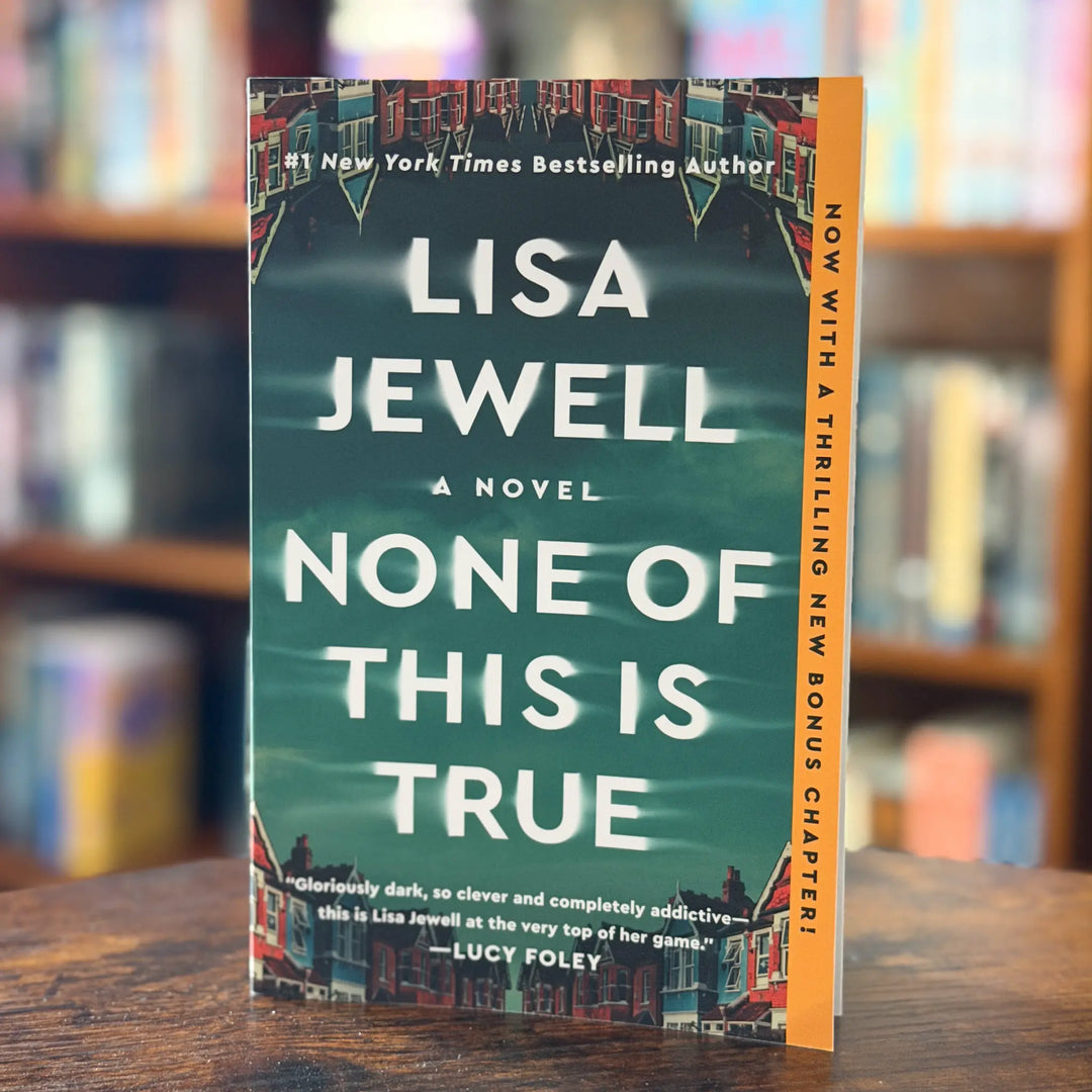 Book titled 'None of This is True' by Lisa Jewell on a wooden surface with a blurred bookshelf background.
