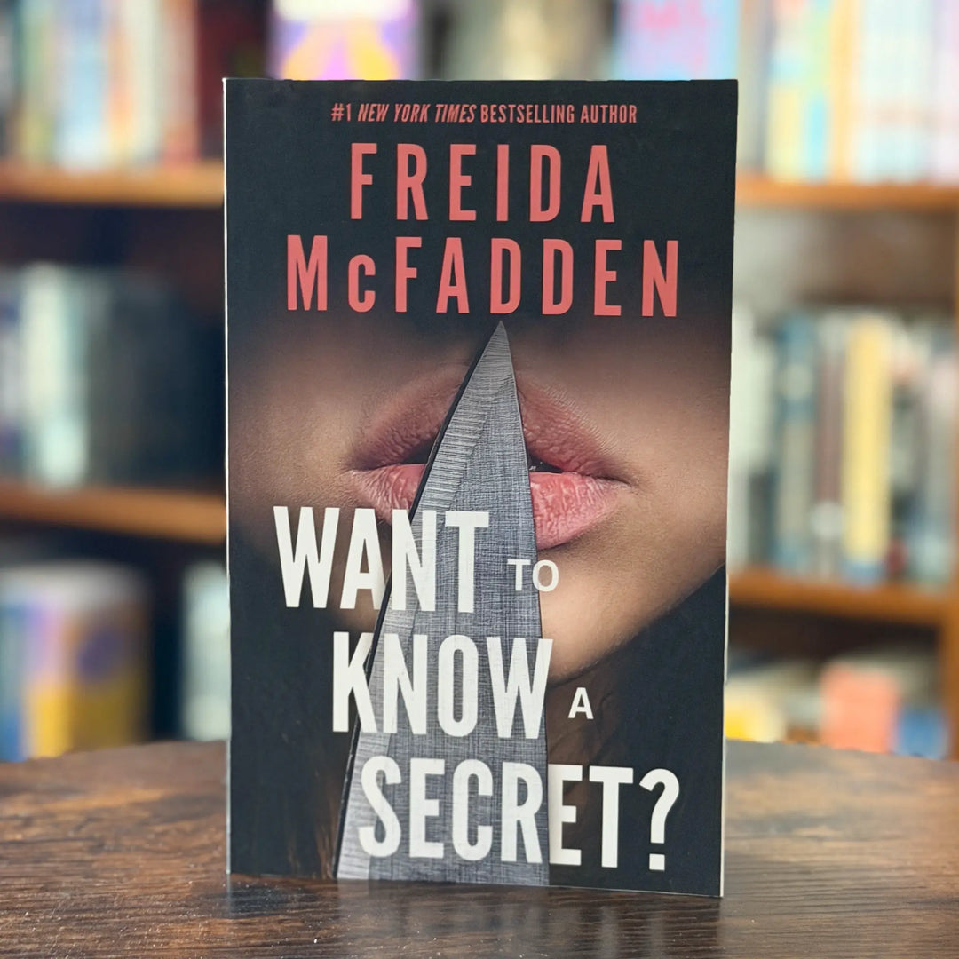 Book cover of 'Want to Know a Secret?' by Freida McFadden on a wooden surface with blurred bookshelf background.