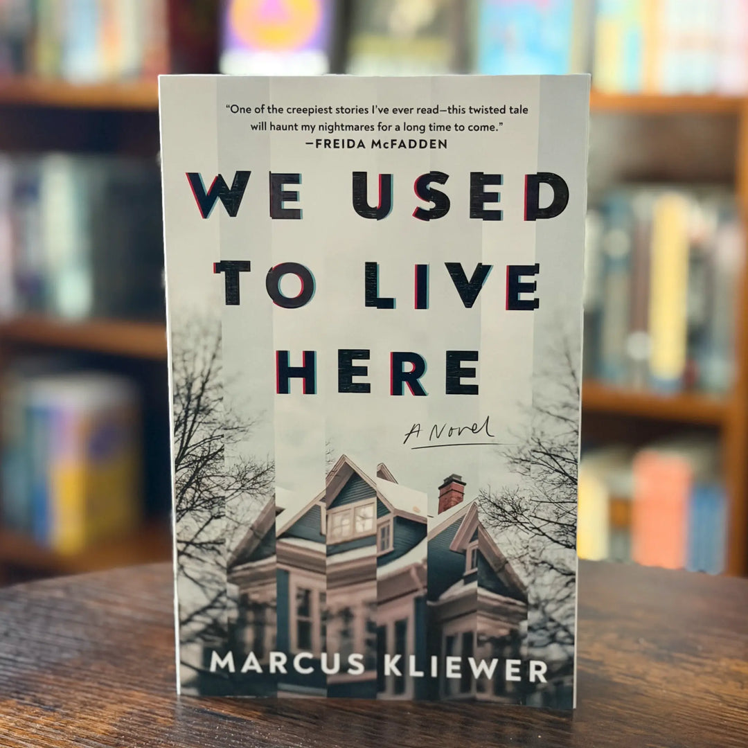 Book titled 'We Used to Live Here' by Marcus Kliewer on a wooden surface with a blurred bookshelf background.
