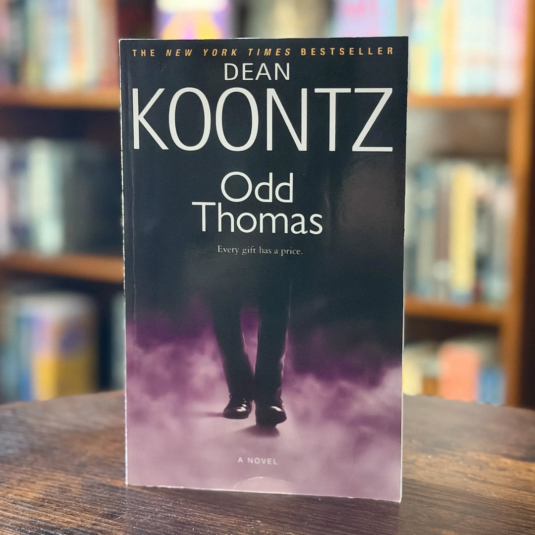 Book titled 'Odd Thomas' by Dean Koontz on a wooden surface with a blurred bookshelf background