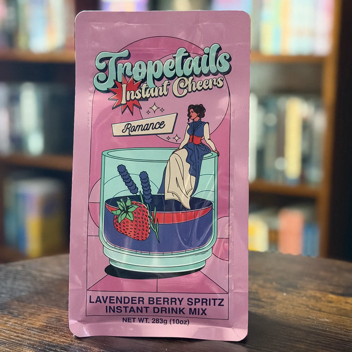 Pink package of Tropetails Instant Cheers drink mix on a wooden surface with a blurred bookshelf background.
