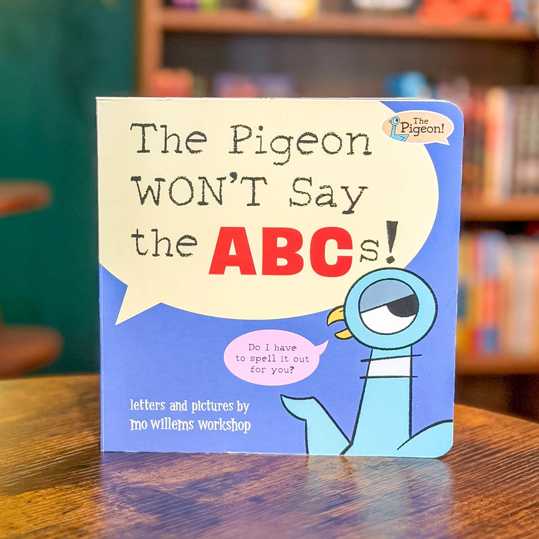 Book titled 'The Pigeon WON'T Say the ABCs!' on a wooden surface with a blurred bookshelf background.