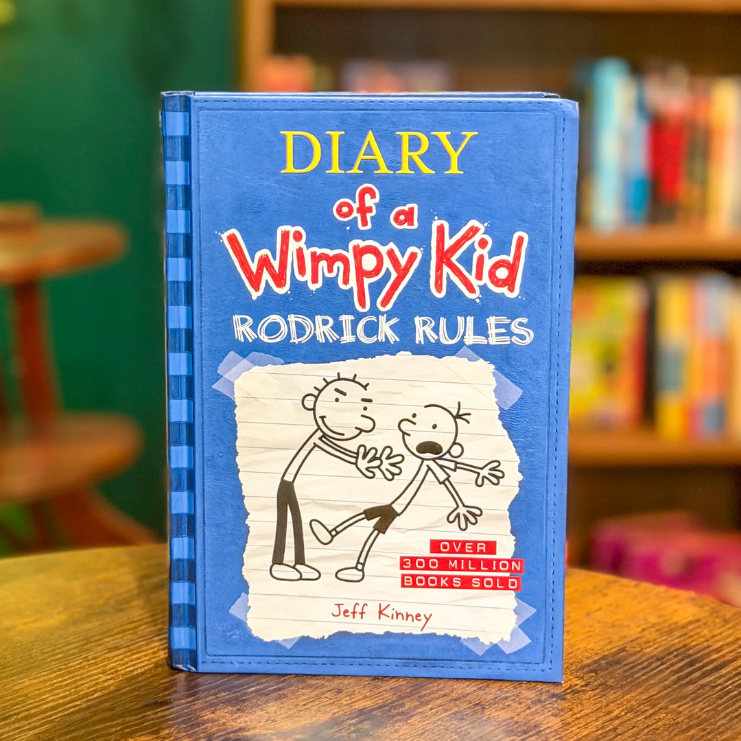 Book titled 'Diary of a Wimpy Kid: Rodrick Rules' on a wooden surface with a blurred bookshelf in the background.