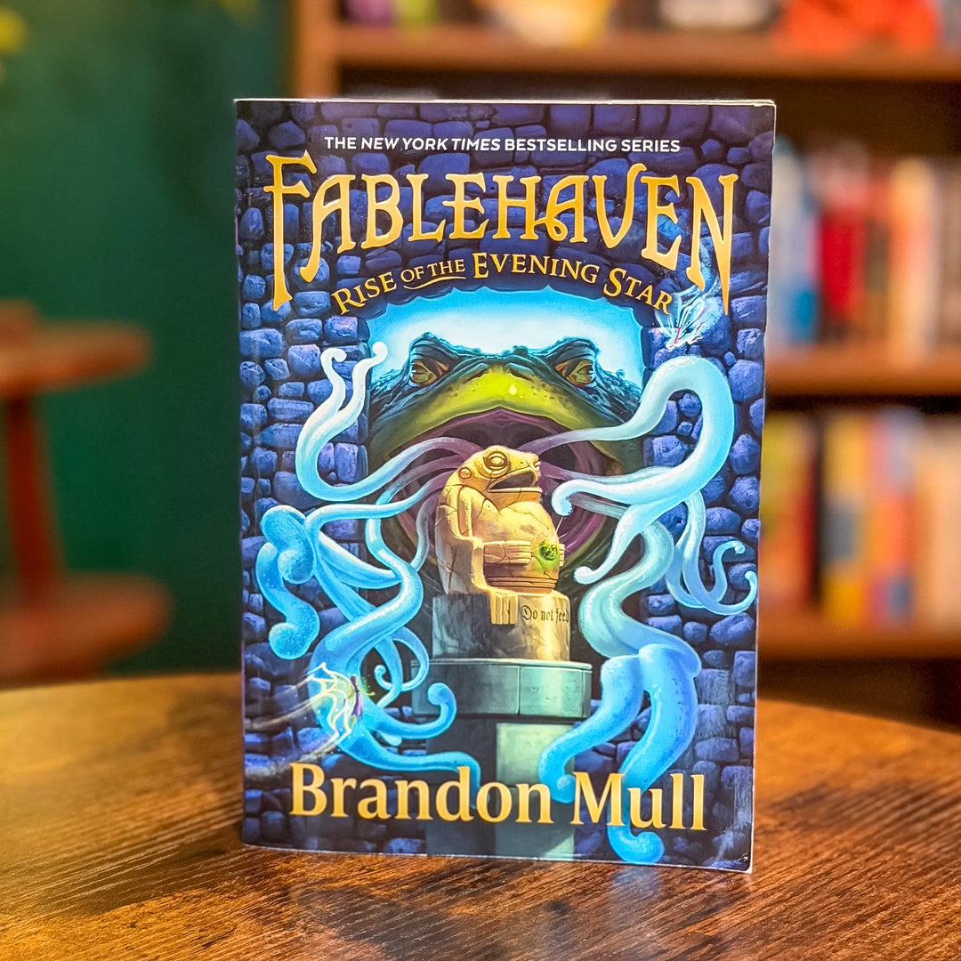 Book titled 'Fablehaven: Rise of the Evening Star' by Brandon Mull on a wooden table with a blurred bookshelf background.