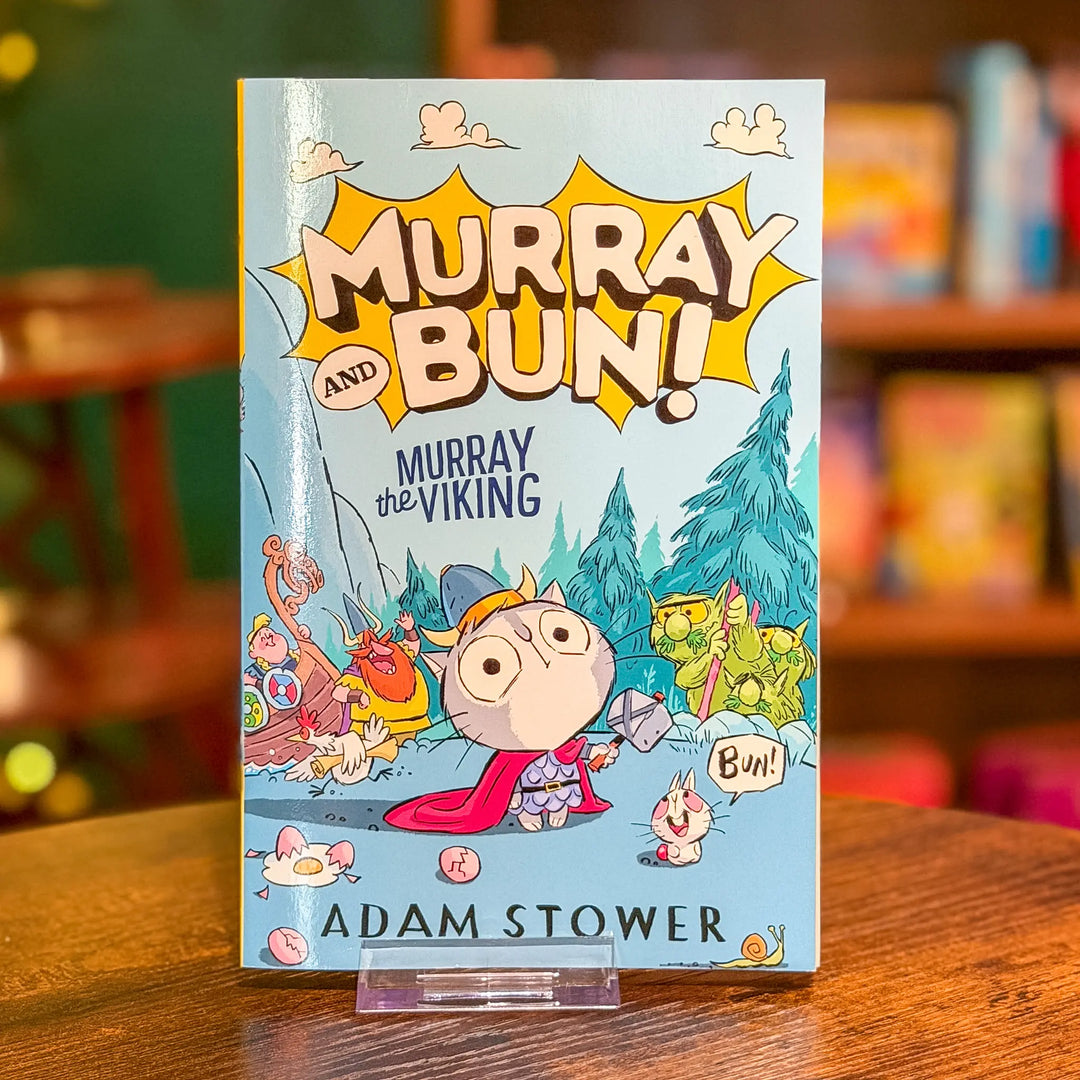 Book titled 'Murray and Bun! Murray the Viking' by Adam Stower on a wooden surface with a blurred bookshelf background.