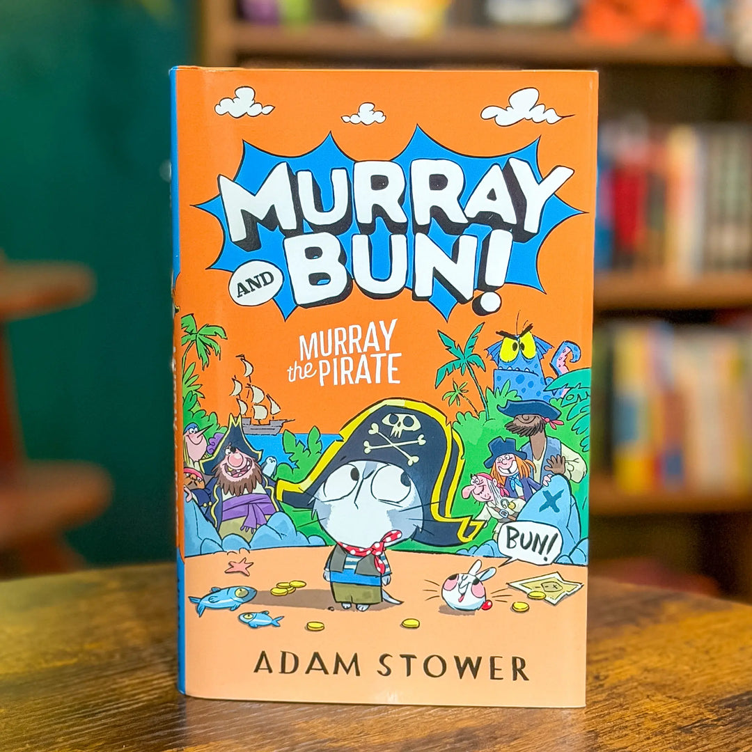 Book titled 'Murray and Bun! Murray the Pirate' by Adam Stower on a wooden surface with a blurred bookshelf background.