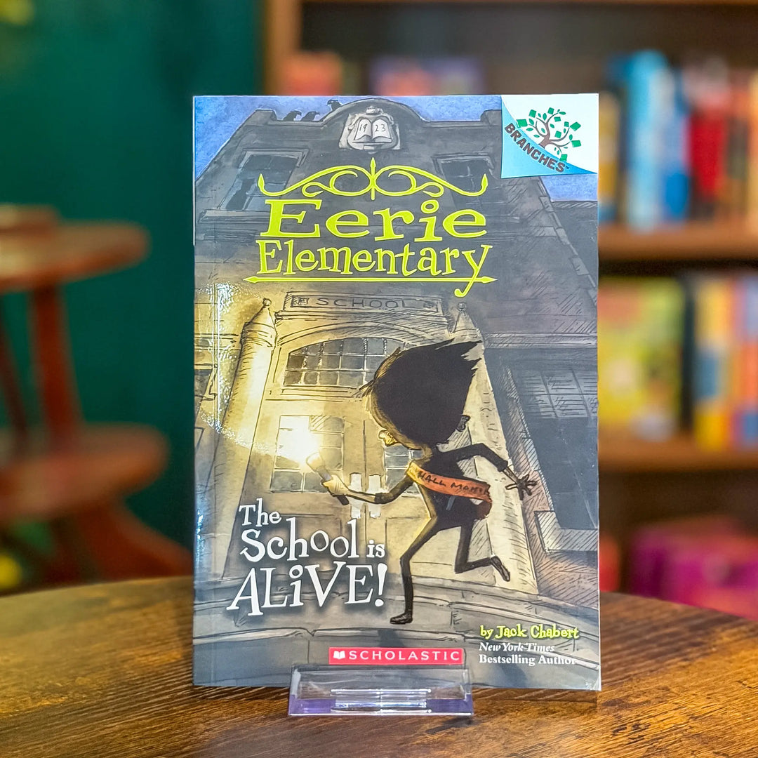 Book titled 'Eerie Elementary: The School is Alive!' by Scholastic on a wooden surface with a blurred bookshelf background.