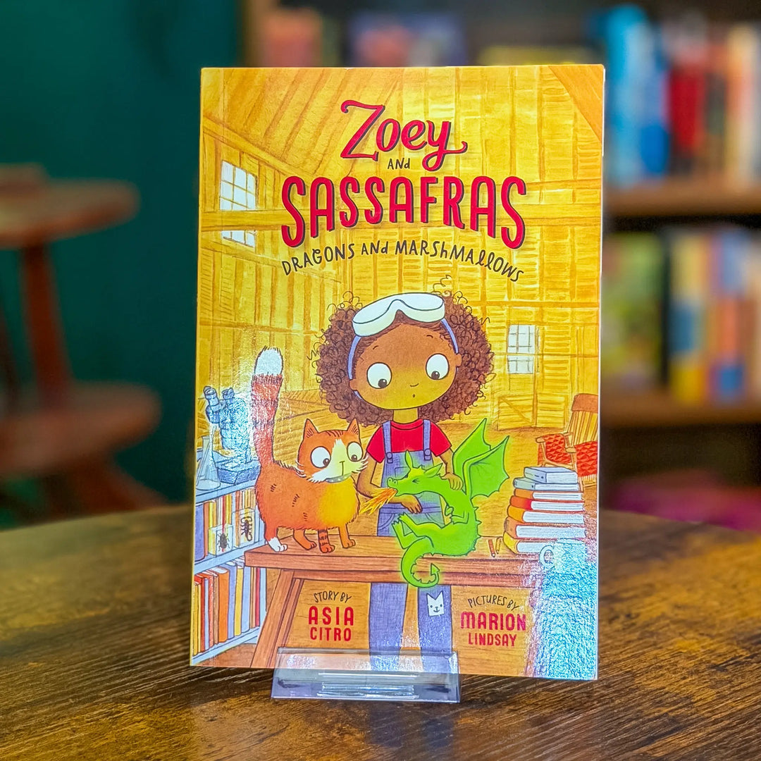 Book cover of 'Zoey and Sassafras: Dragons and Marshmallows' on a wooden surface with a blurred bookshelf background.