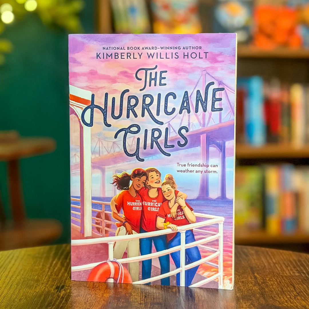 Book titled 'The Hurricane Girls' by Kimberly Willis Holt on a table with a blurred bookshelf background.