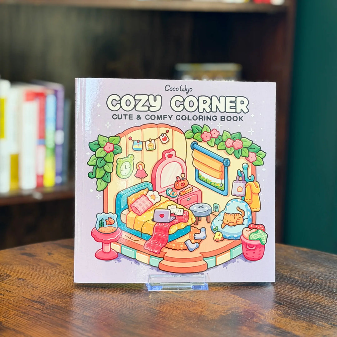Coloring book titled 'Cozy Corner' by Coco Wyo on a wooden surface with a blurred bookshelf in the background.