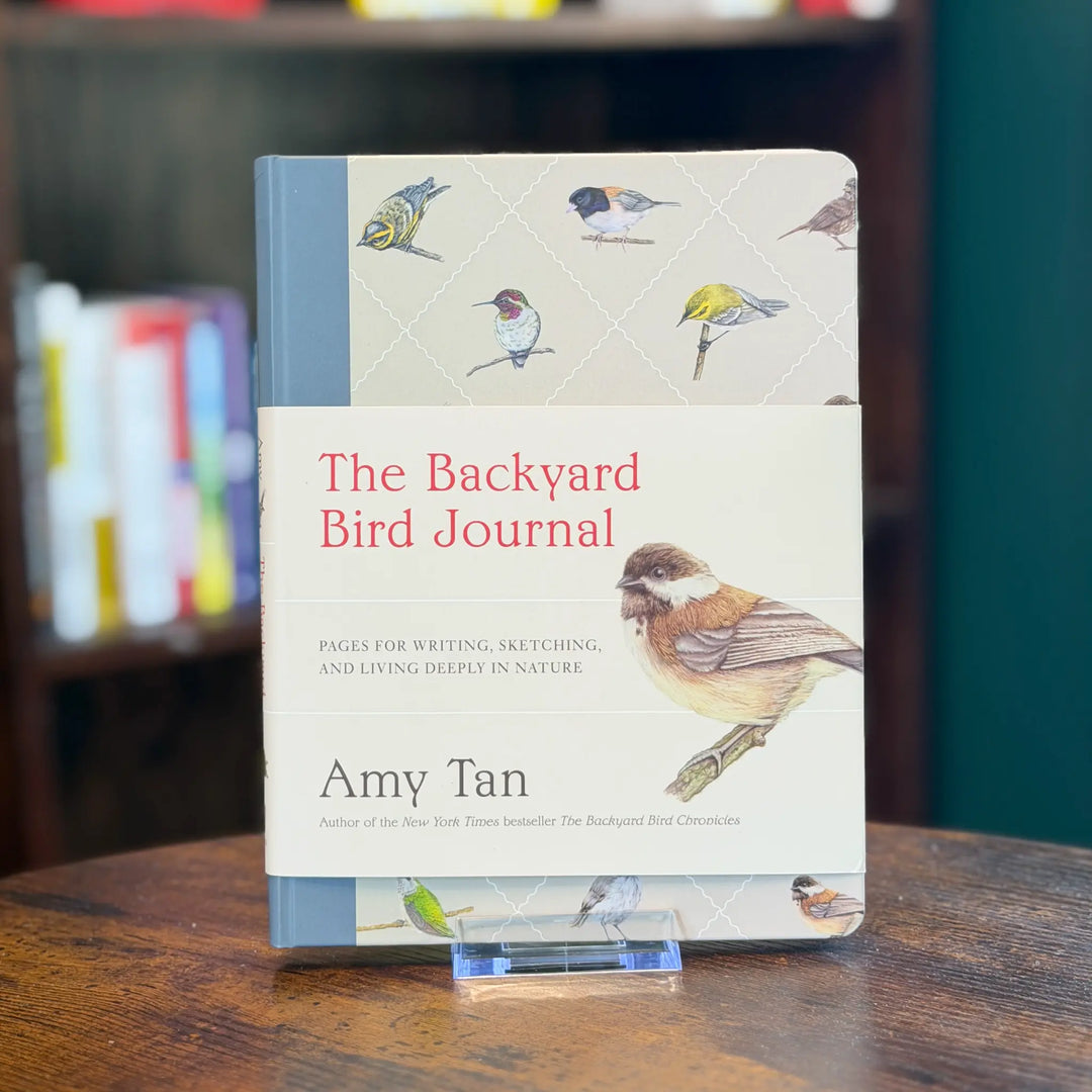 Book titled 'The Backyard Bird Journal' by Amy Tan on a wooden surface with a blurred bookshelf background.