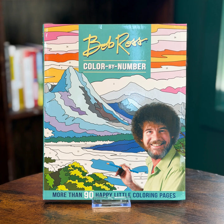 Bob Ross color-by-number coloring book on a wooden surface with a blurred background