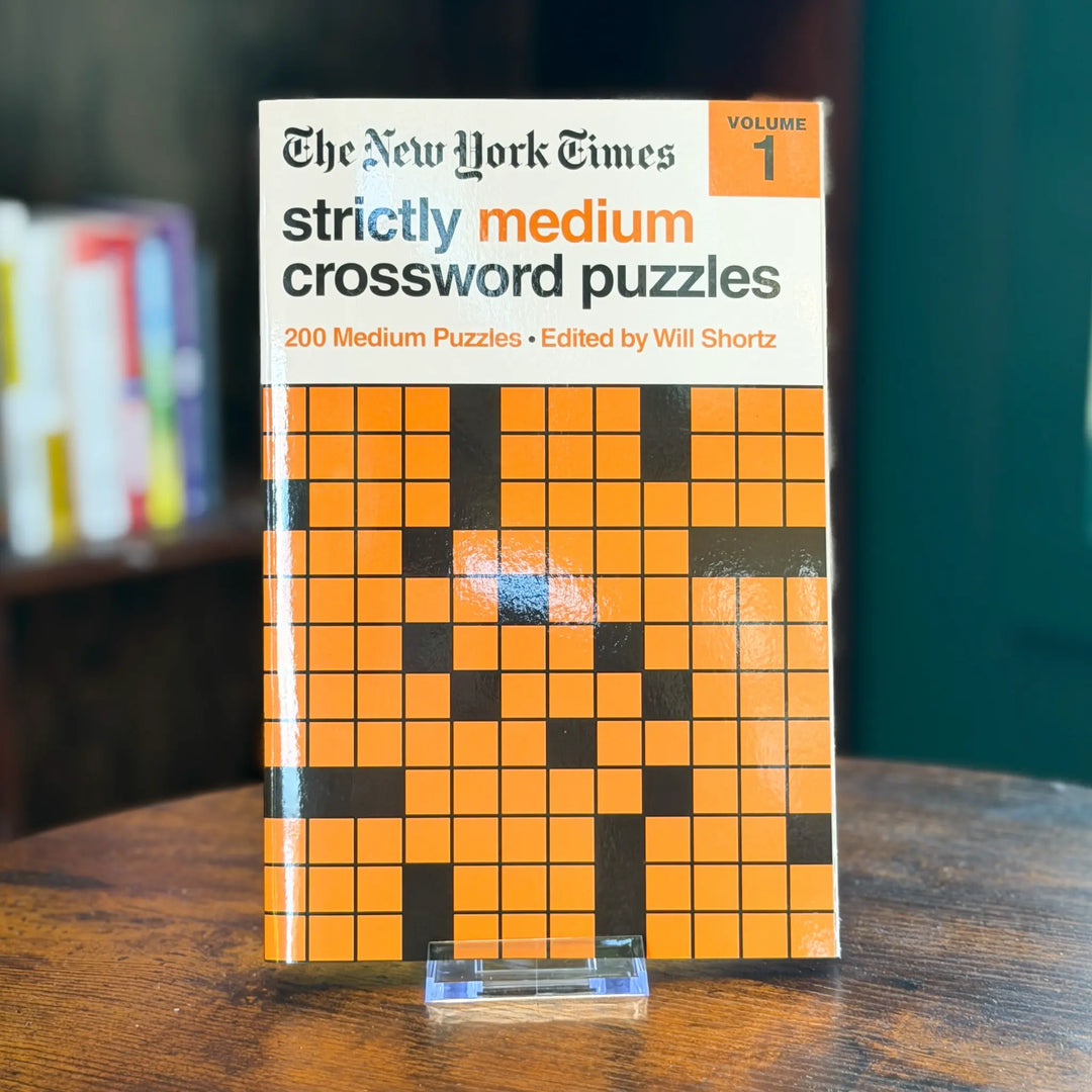 Book titled 'The New York Times Strictly Medium Crossword Puzzles' on a wooden surface with a blurred bookshelf in the background.