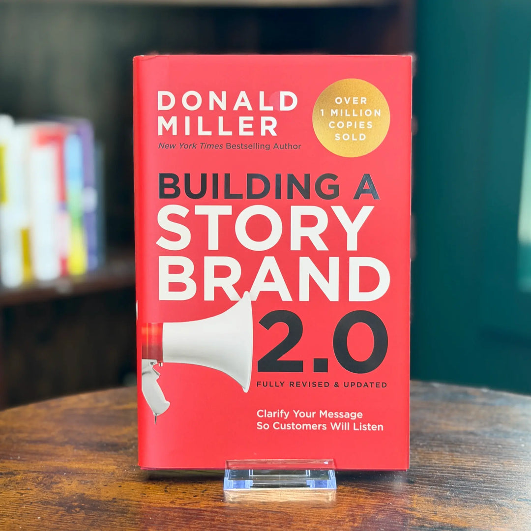 Book titled 'Building a Story Brand 2.0' by Donald Miller on a wooden surface with a blurred bookshelf in the background.