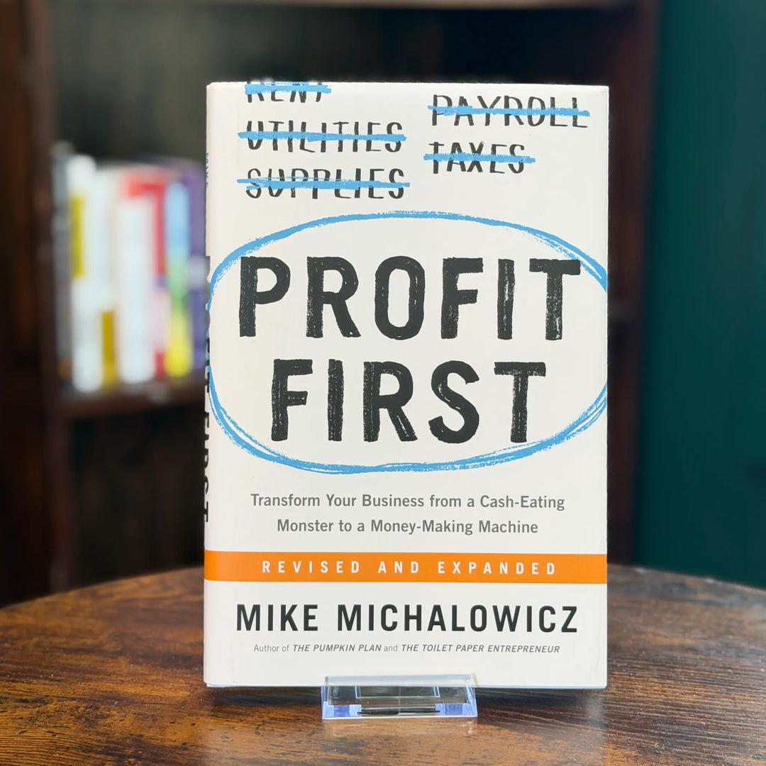 Book titled 'Profit First' by Mike Michalowicz on a wooden surface with blurred books in the background.