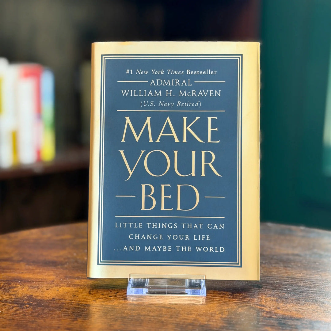 Book titled 'Make Your Bed' by William H. McRaven on a wooden surface with blurred books in the background.