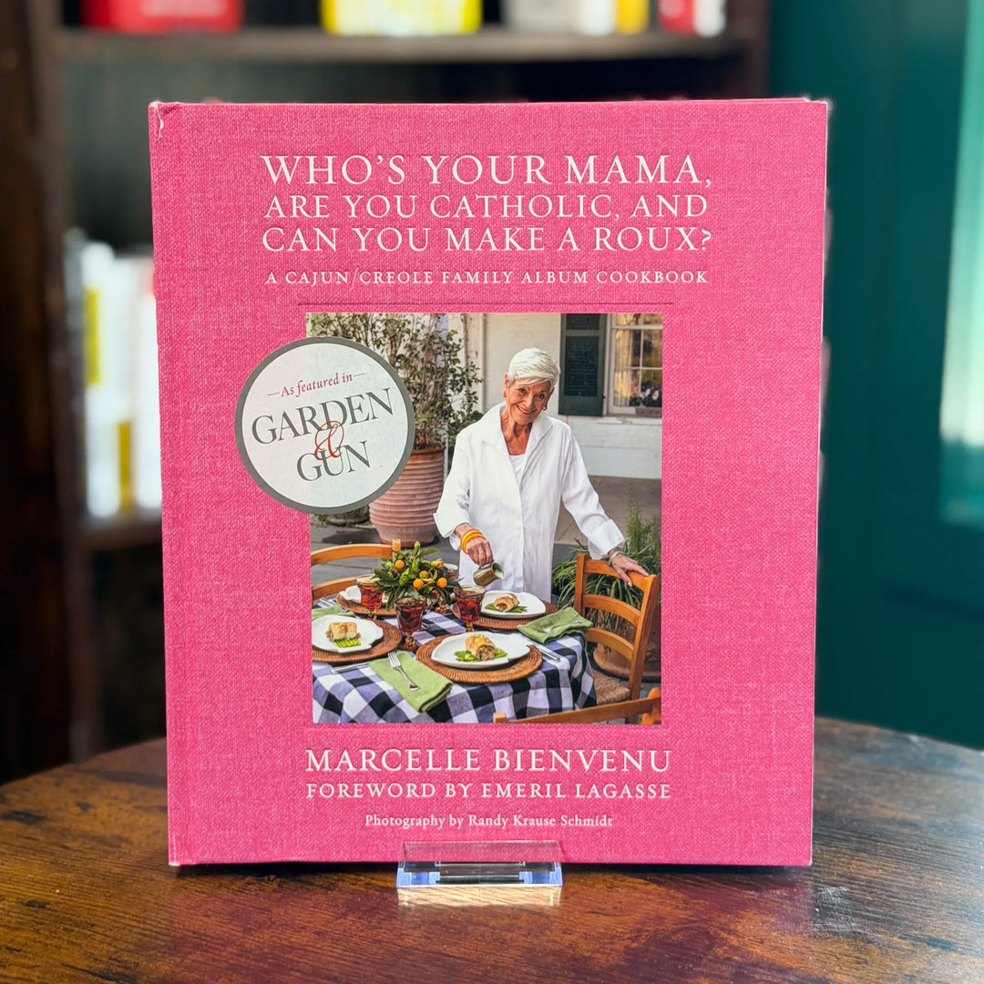 Pink cookbook titled 'Who's Your Mama, Are You Catholic, and Can You Make a Roux?' on a wooden surface.
