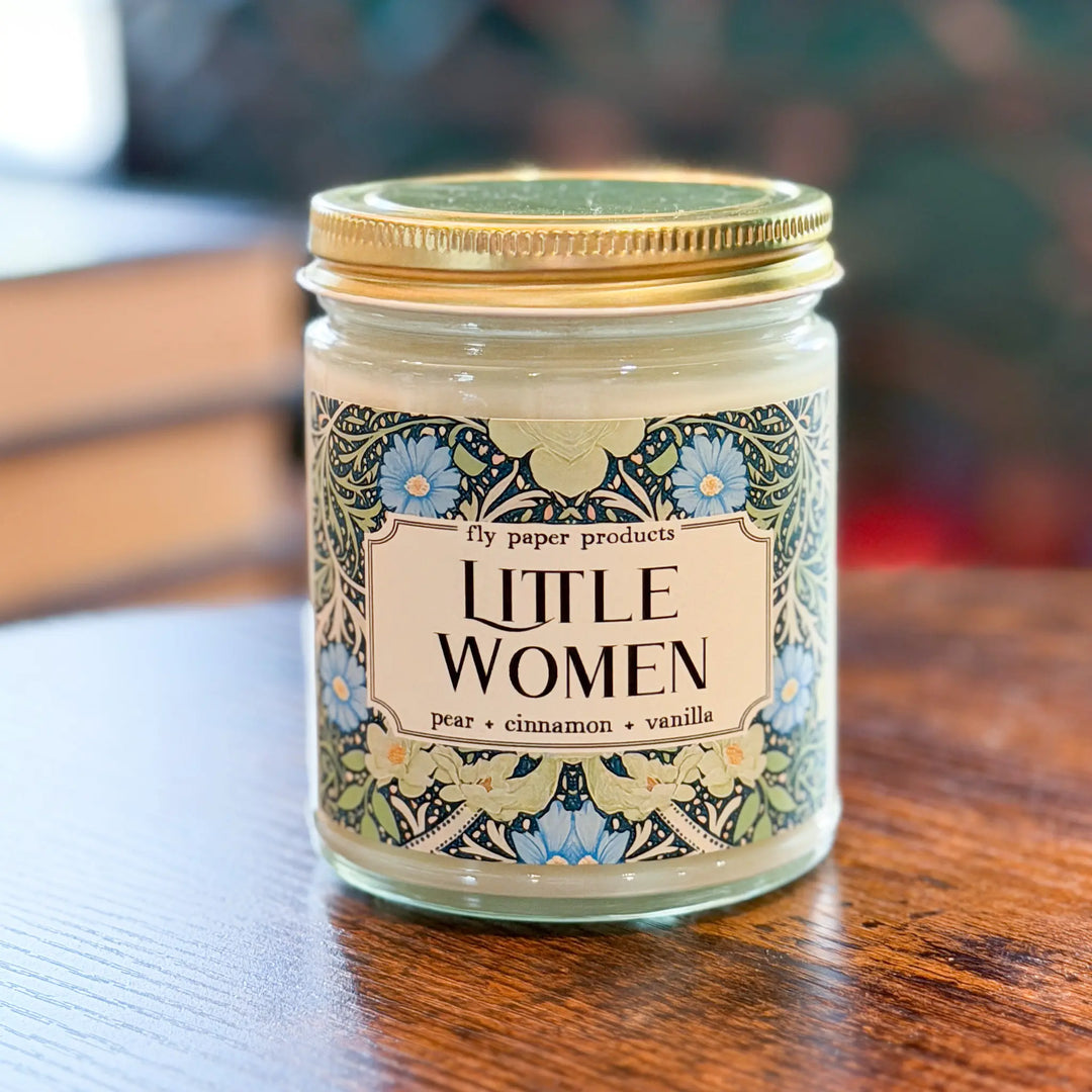 Candle jar labeled 'Little Women' on a wooden surface with a blurred background