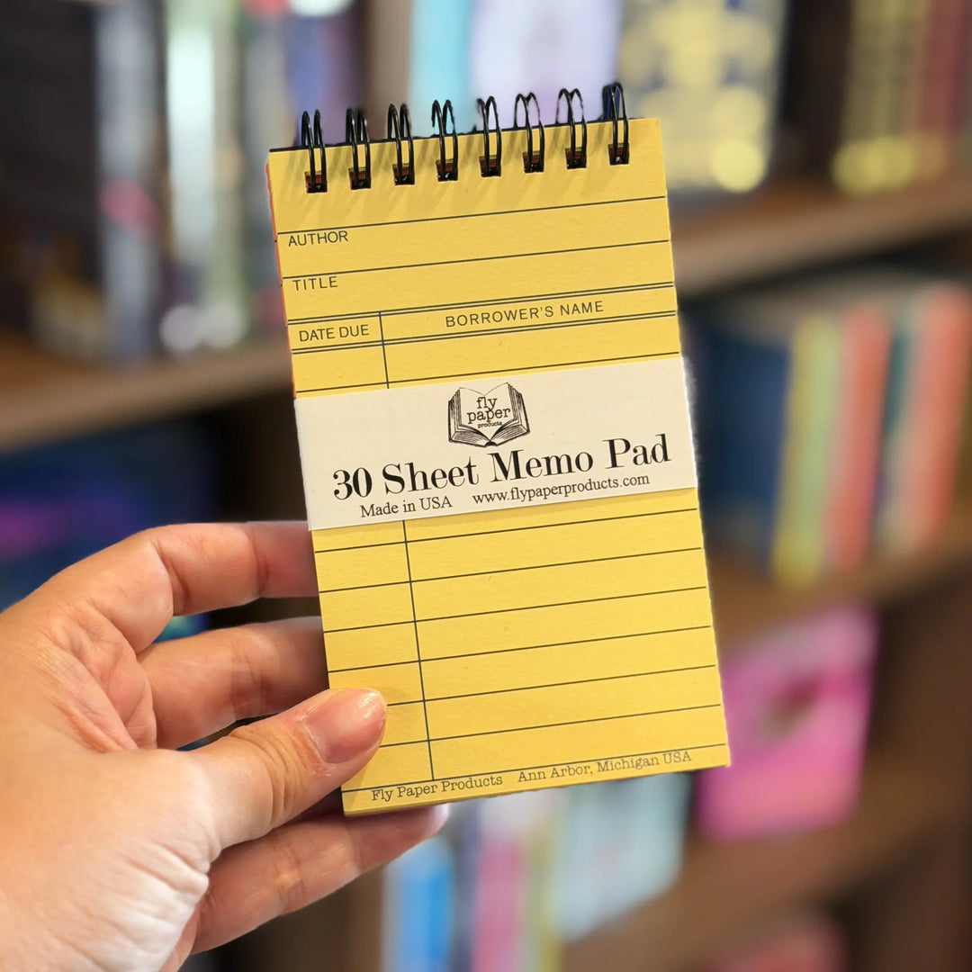 Yellow memo pad with black text held by a hand against a blurred bookshelf background