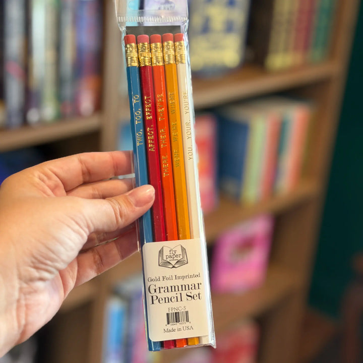 Packaged set of colored pencils held by a hand with a blurred bookshelf background