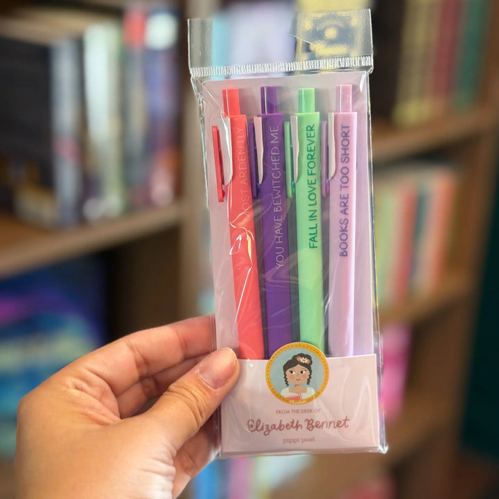 Packaged pens with colorful caps held by a hand in front of a blurred bookshelf.
