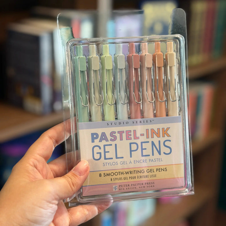 Pack of pastel ink gel pens held by a hand with a blurred bookshelf background