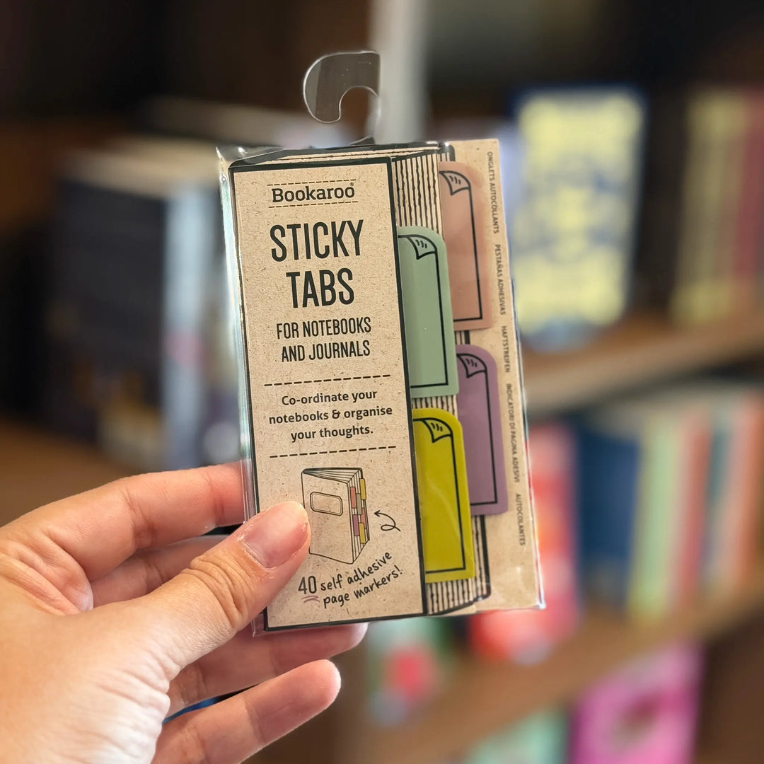 Hand holding a package of Bookaroo sticky tabs for notebooks and journals against a blurred bookshelf background.