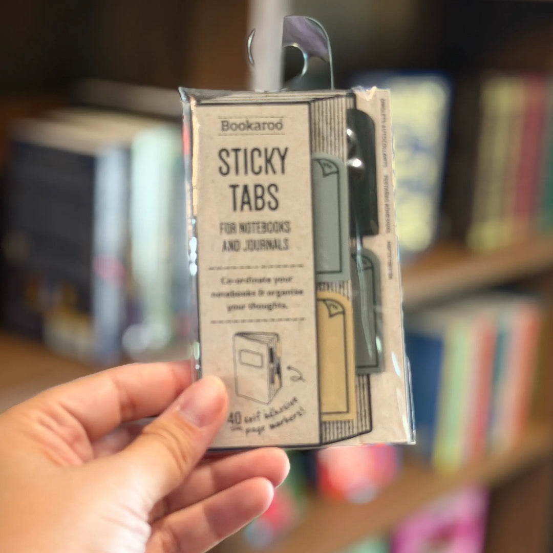 Hand holding a package of Bookaroo sticky tabs with a blurred bookshelf background