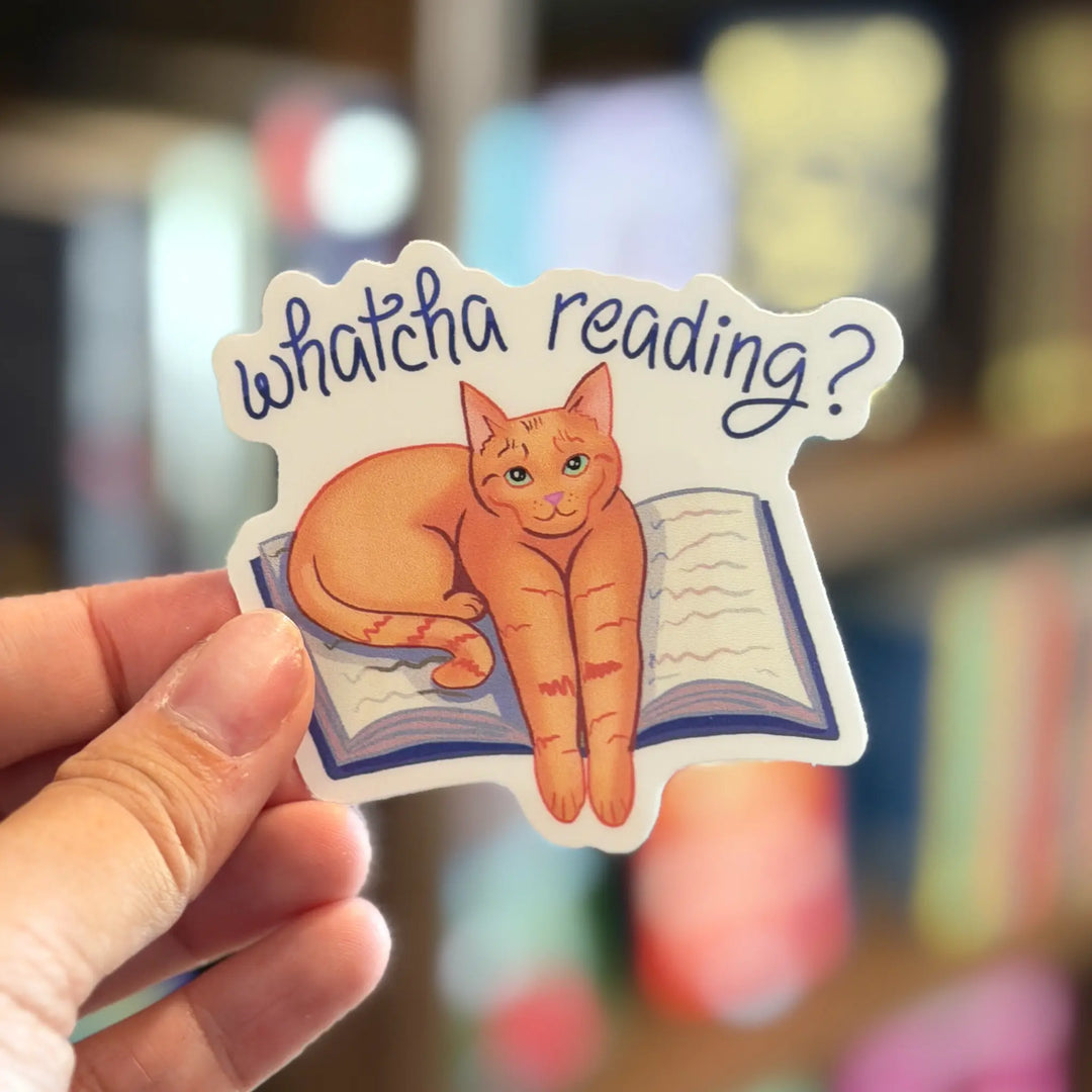 Sticker with a cat sitting on a book and 'whatcha reading?' text, held in front of a blurred bookshelf.