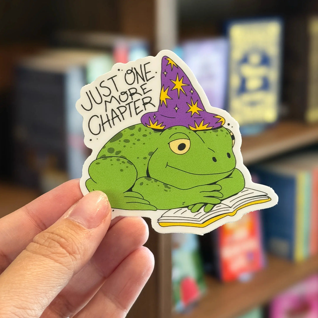 Sticker of a frog wearing a wizard hat with 'Just One More Chapter' text, held in front of a bookshelf.