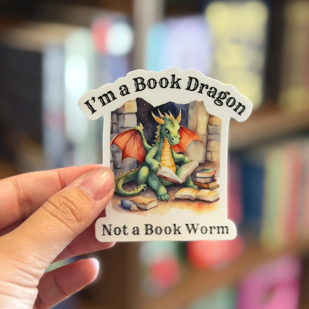 Sticker with a dragon design and text held by a hand against a blurred bookshelf background
