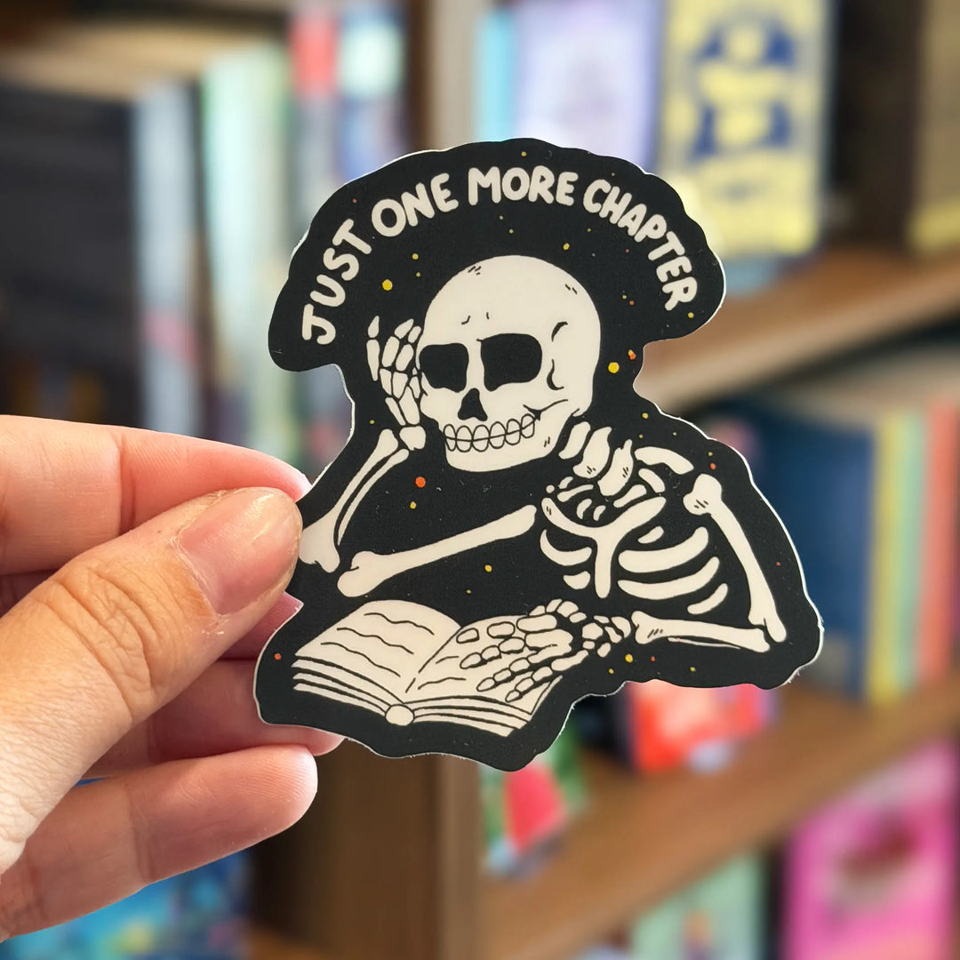 Sticker of a skeleton reading a book with 'Just One More Chapter' text, held in front of a bookshelf.