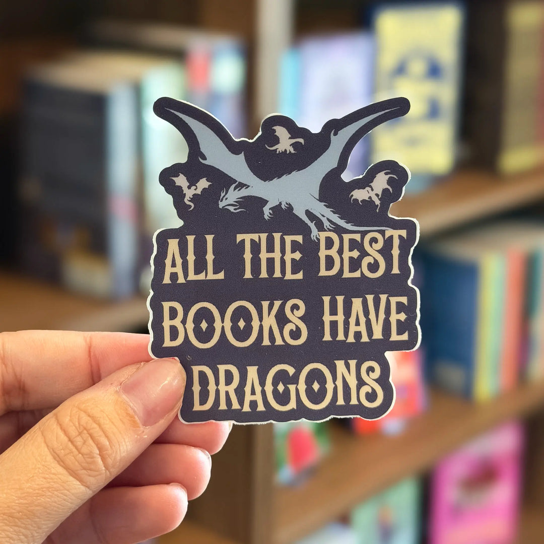 Sticker with 'All the best books have dragons' text held in front of a bookshelf.