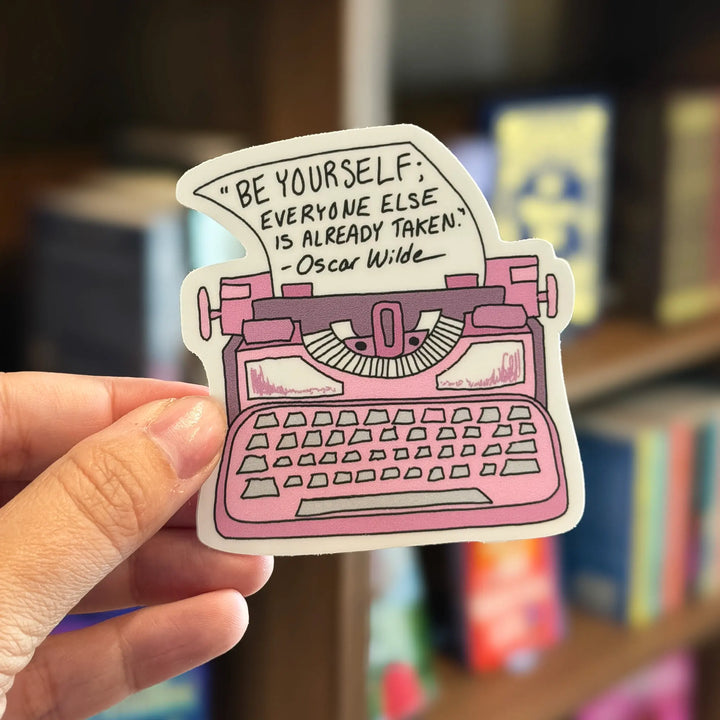 Pink typewriter sticker with Oscar Wilde quote held by a hand in front of a bookshelf.