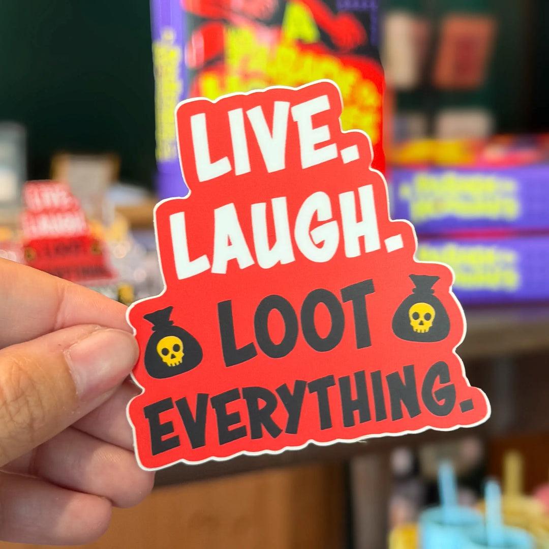 Sticker with 'Live. Laugh. Loot. Everything.' text held by a hand in front of a blurred store background.
