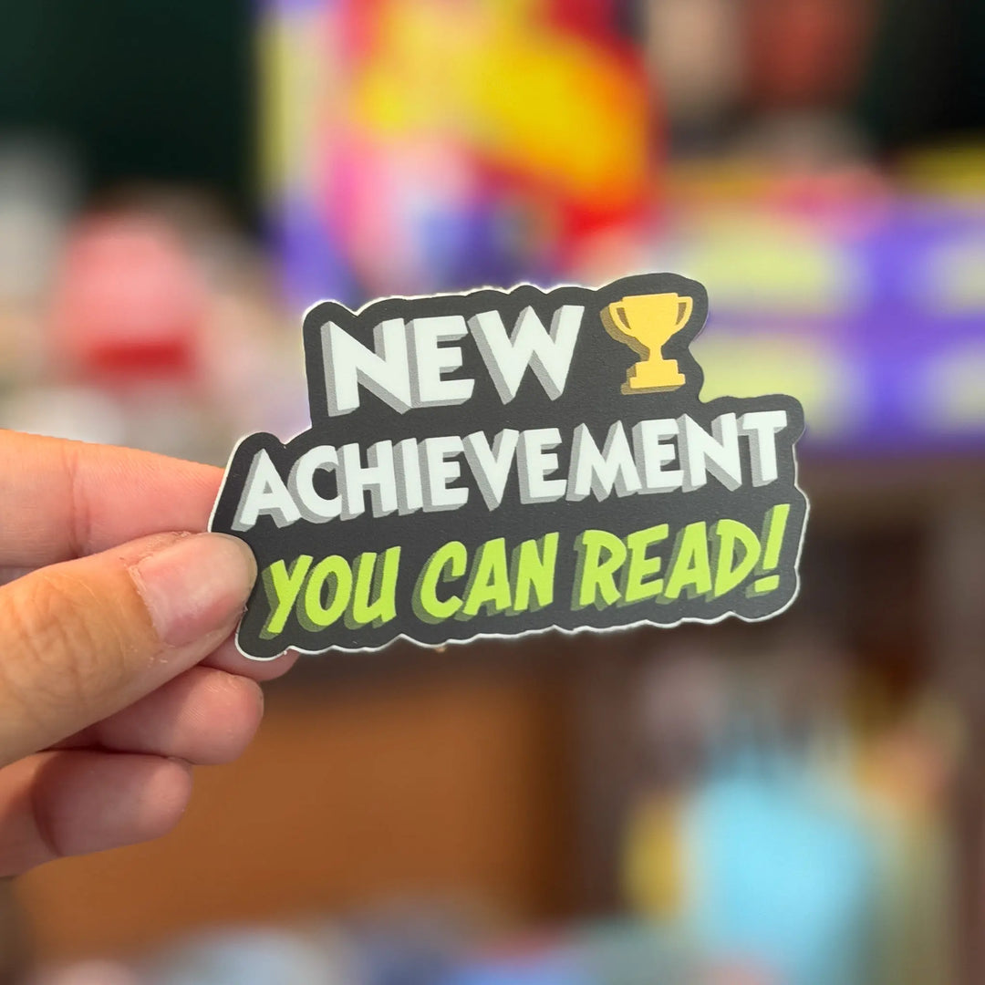 Hand holding a sticker with 'New Achievement You Can Read!' text against a colorful blurred background
