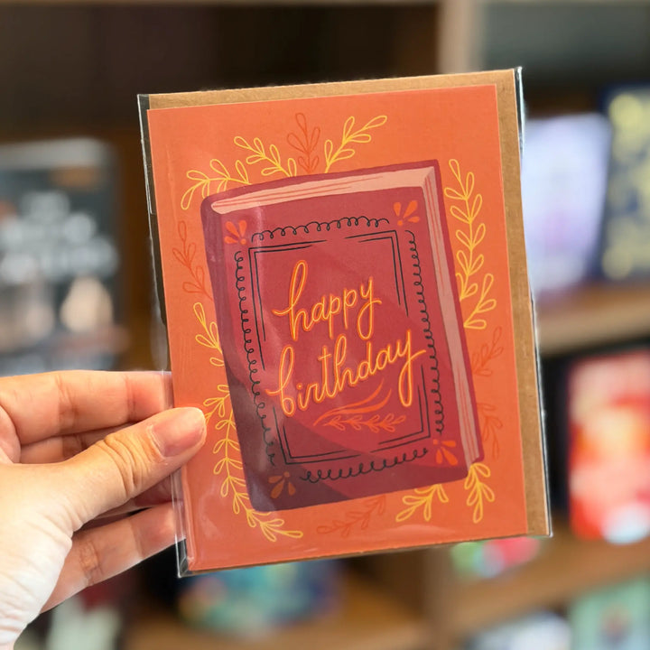 Hand holding a birthday card with decorative text and border. Text reads &