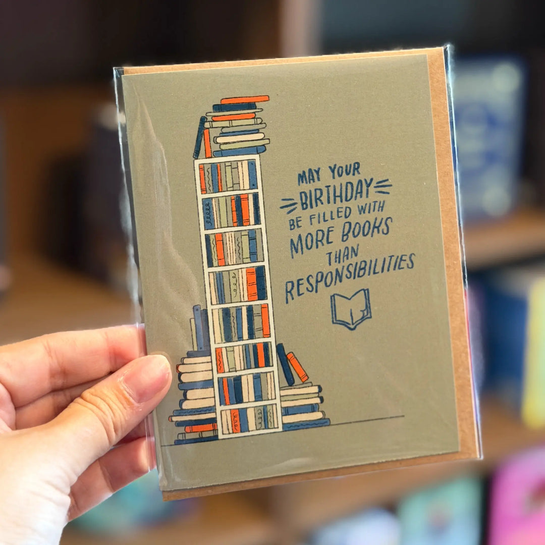 Greeting card with bookshelf illustration and birthday message held by a hand. Text reads 'May your birthday be filled with more books than responsibilities'