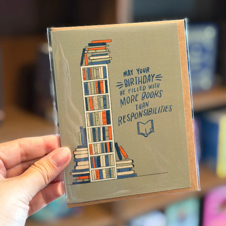 Greeting card with bookshelf illustration and birthday message held by a hand. Text reads &