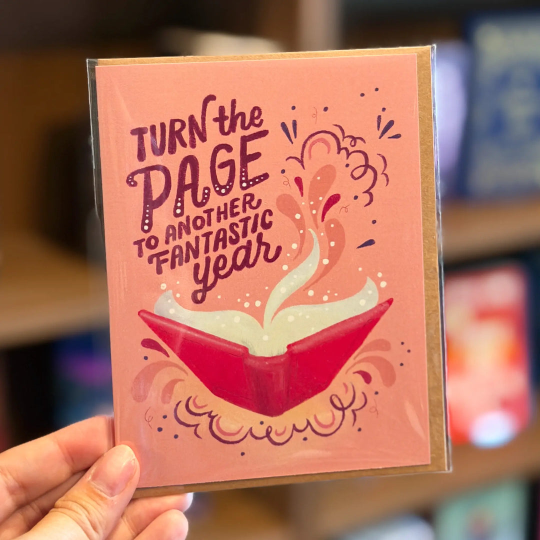 Greeting card with 'Turn the page to another fantastic year' text and book illustration, held in a hand.
