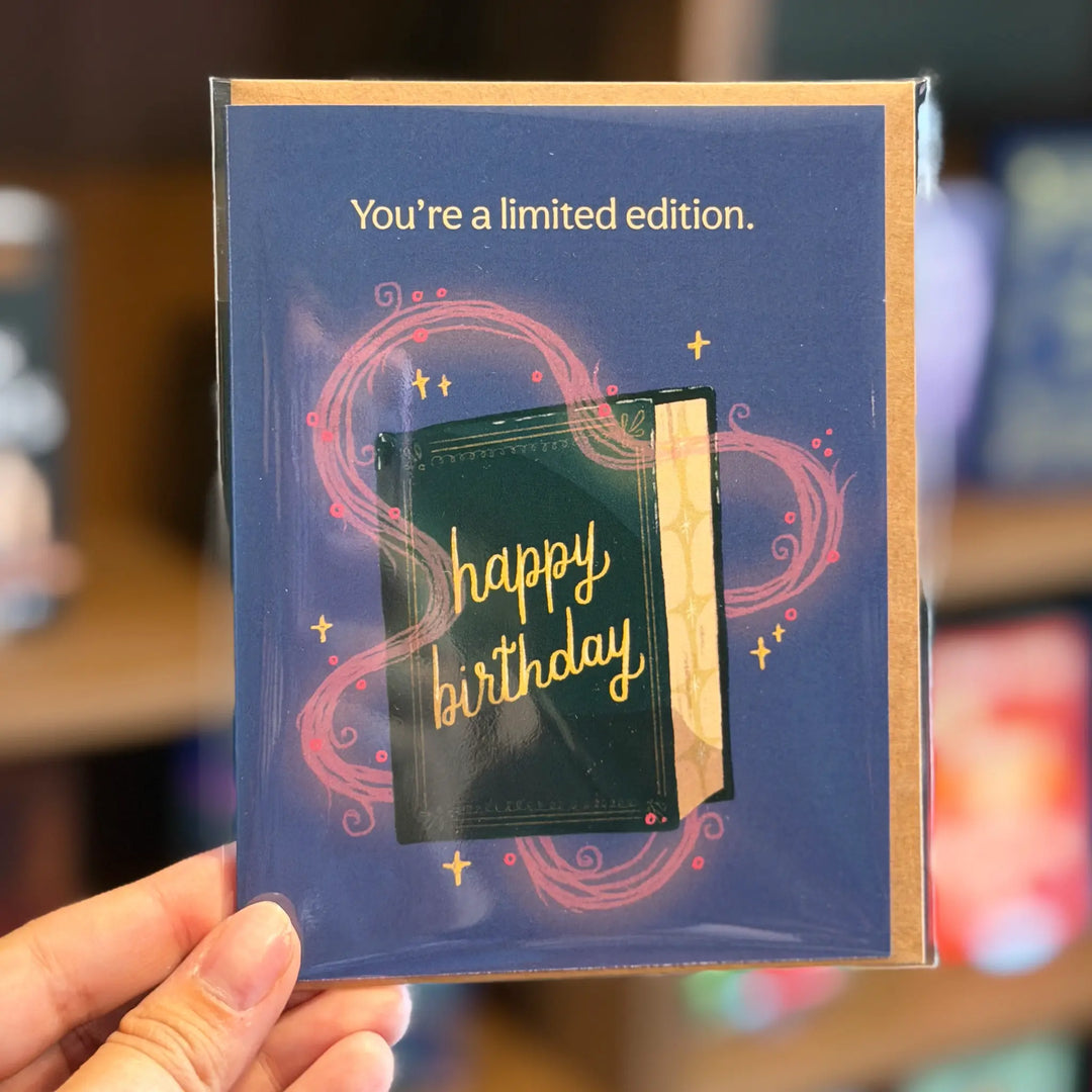 Blue birthday card with text and a small green book-shaped object, held by a hand. Text reads 'You're a limited edition' and 'happy birthday'