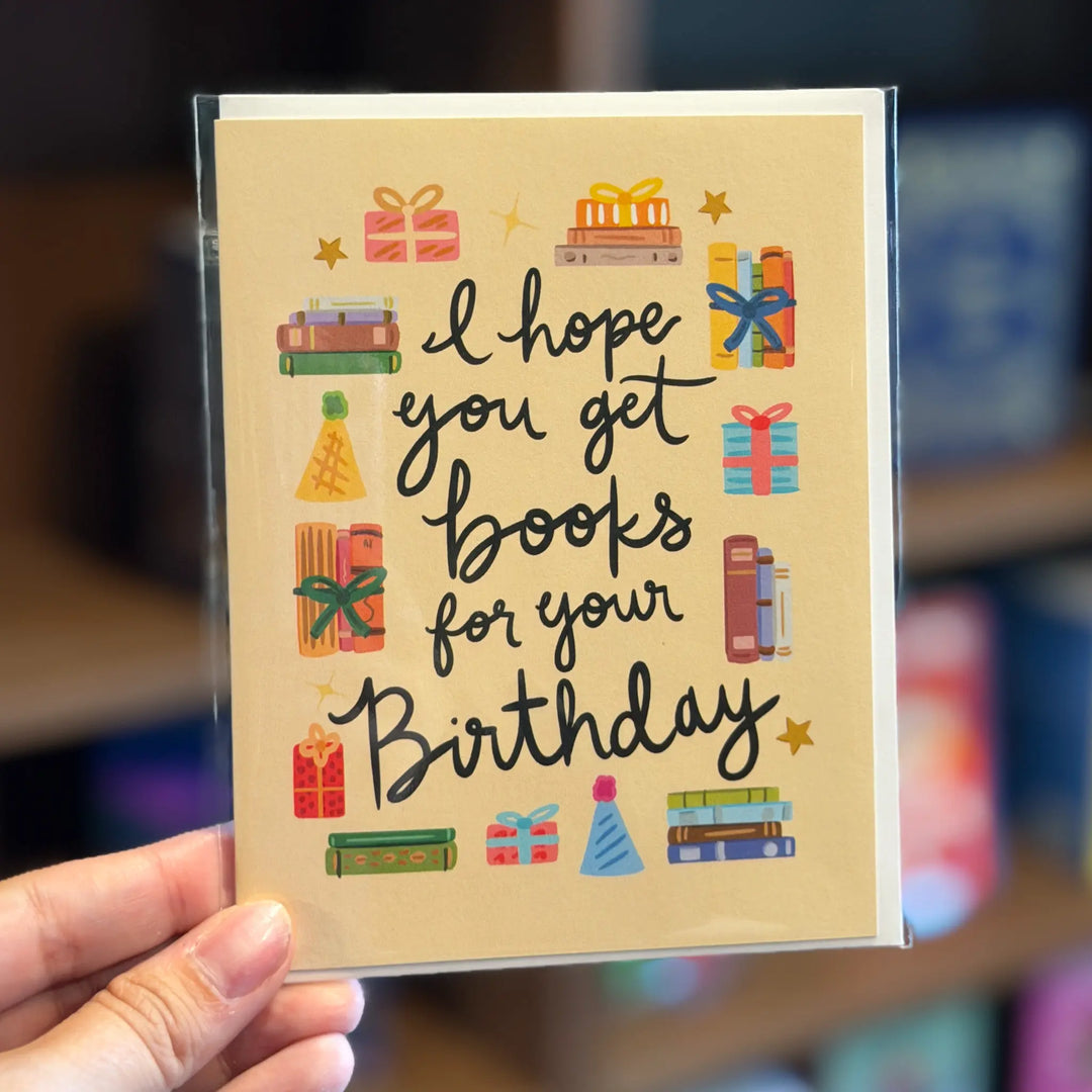 Hand holding a birthday card with book-themed illustrations and text. Text reads 'I hope you get books for your birthday'