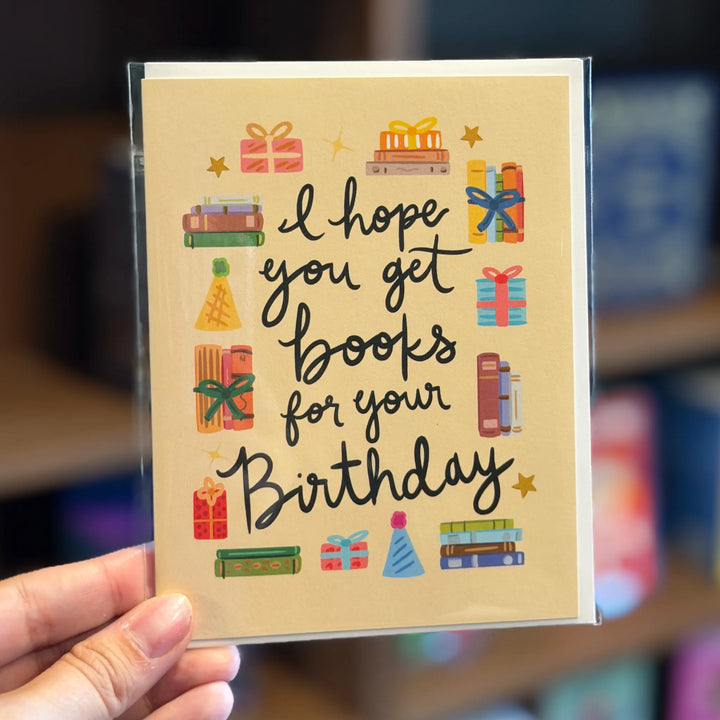 Hand holding a birthday card with book-themed illustrations and text. Text reads &
