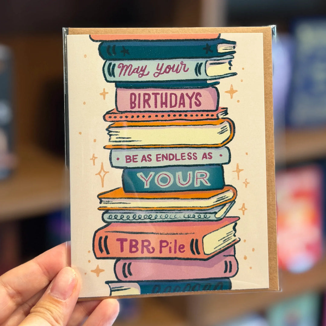 Greeting card with a stack of books and text held by a hand, with a blurred bookshelf background. Text reads 'May your birthdays be as endless as your TBR pile'
