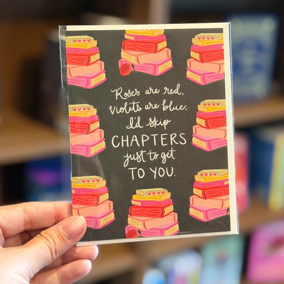 Card with book stack design and text held by a hand, blurred background. Text reads 'Roses are red, violets are blue, I'd skip chapters just to get to you'