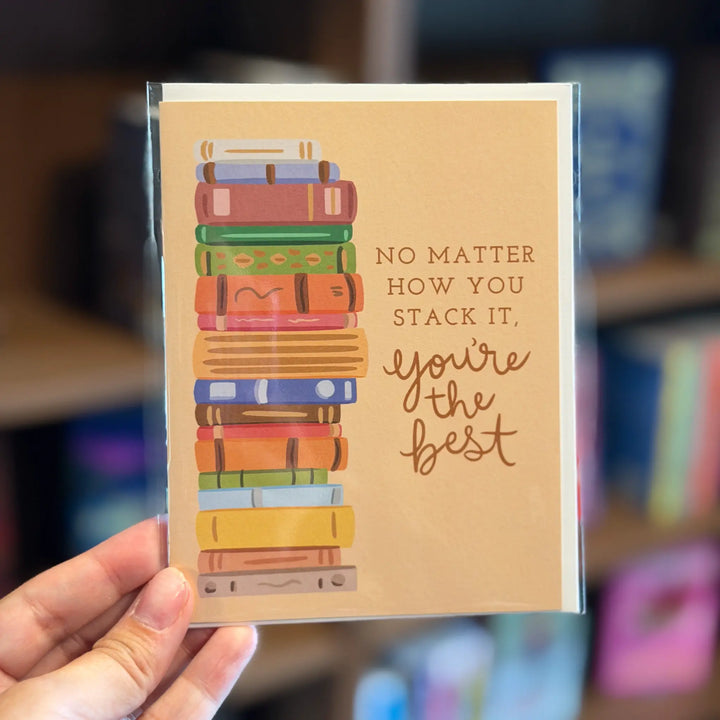 Card with colorful book stack illustration and text held by a hand in front of a blurred bookshelf background. Text reads &
