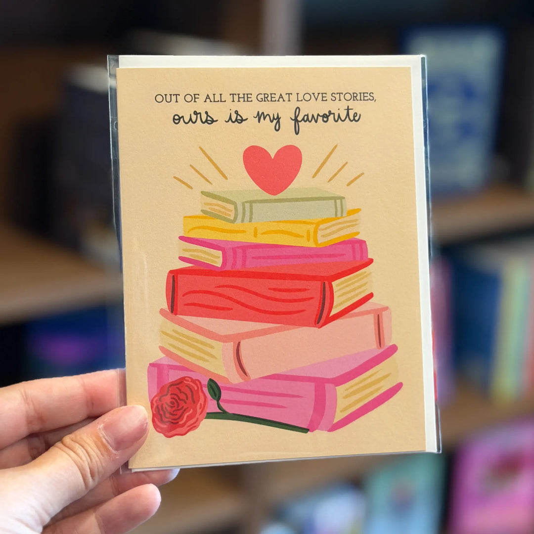Card with books and heart design held by a hand, text 'Out of all the great love stories, ours is my favorite' on a blurred background.