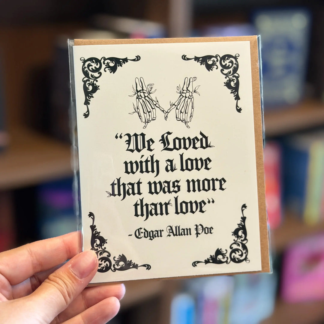 Card with skeleton hands and Edgar Allan Poe quote held by a hand against a blurred bookshelf background. Text reads 'We loved with a love that was more than love'