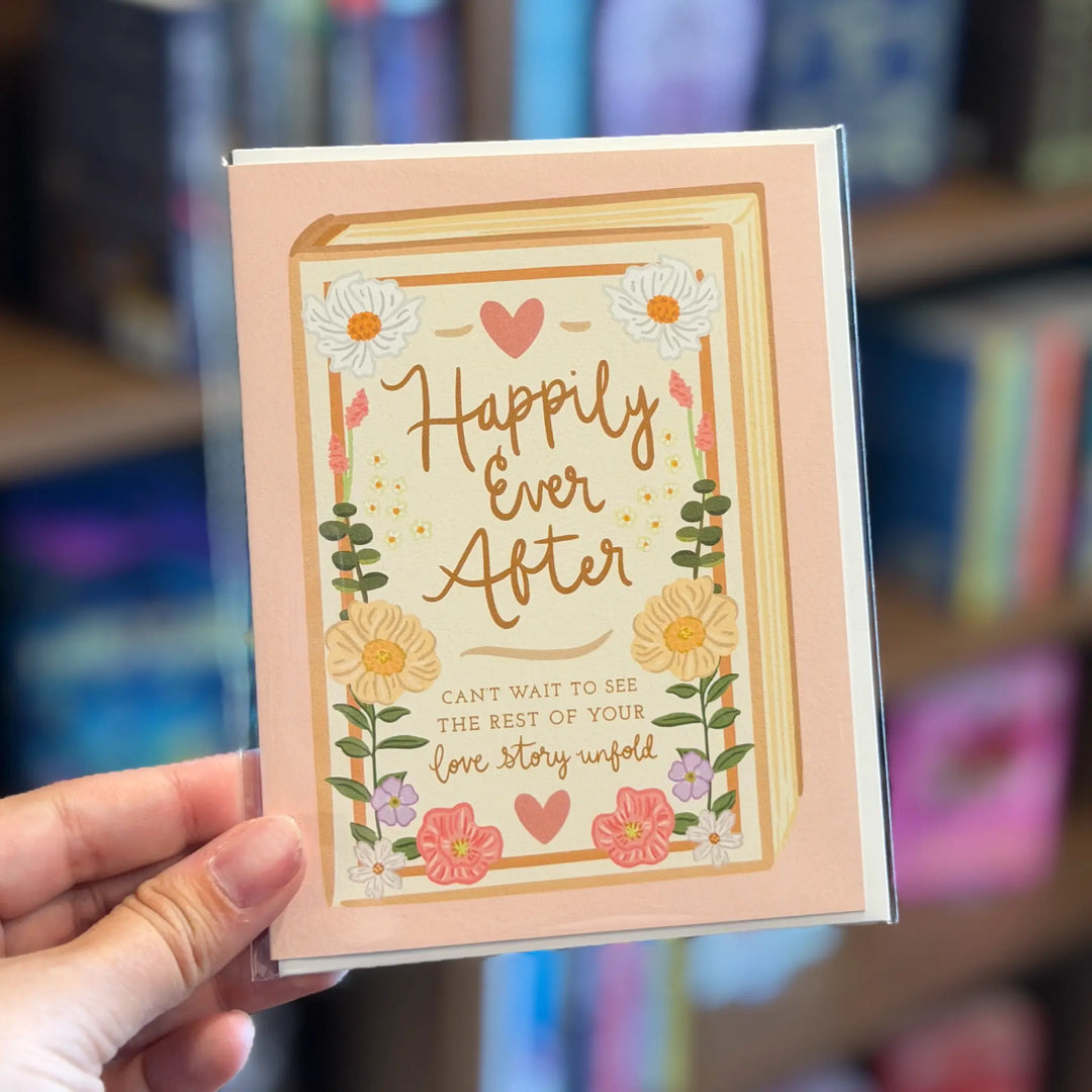 Card with 'Happily Ever After' text held by a hand against a blurred bookshelf background. Text below reads 'Can't wait to see the rest of your love story unfold'