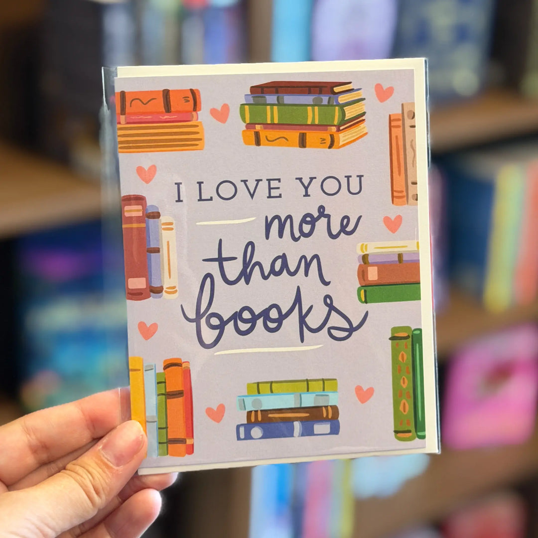 Card with 'I love you more than books' text held by a hand in a blurred bookstore setting